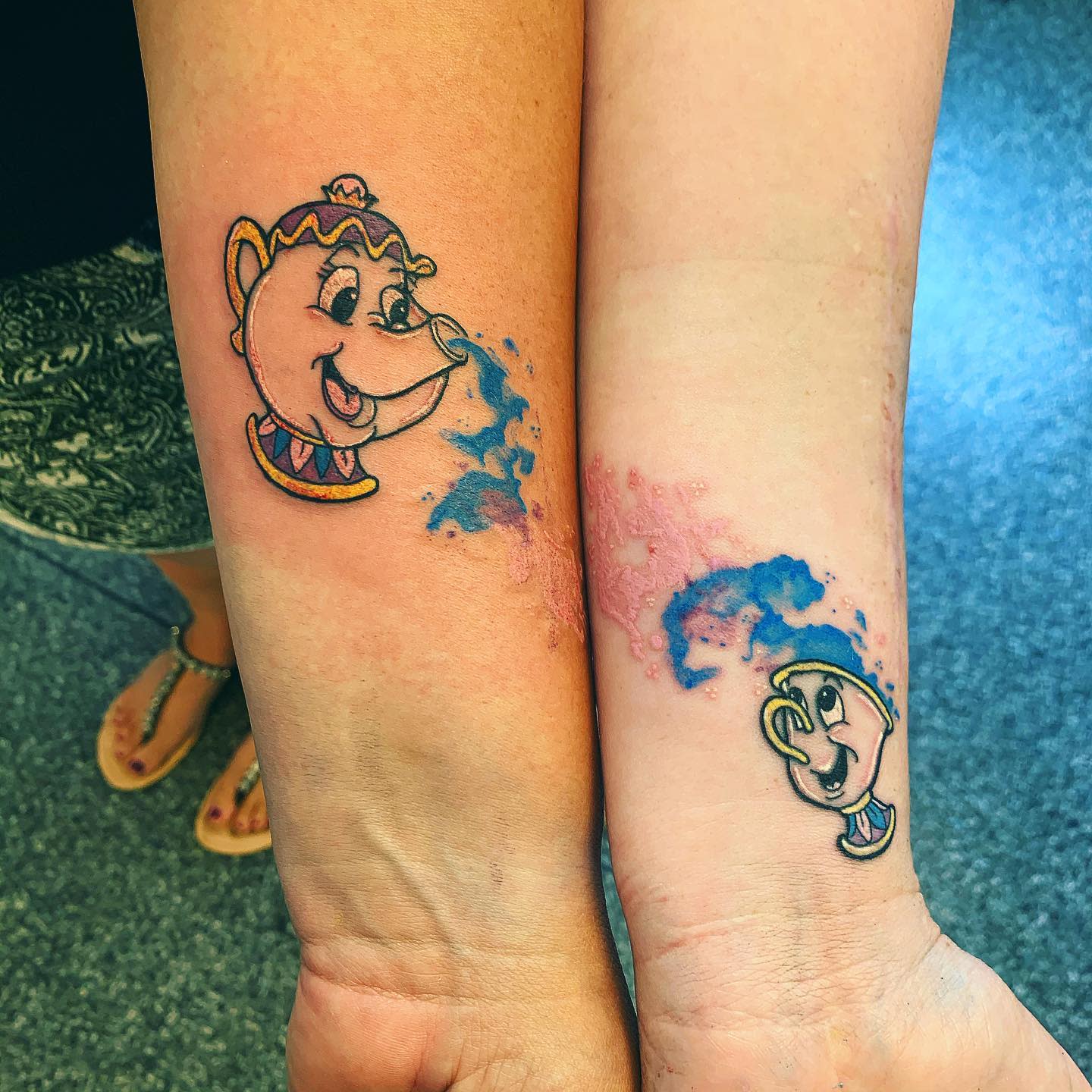 Two forearms with matching teapot and teacup tattoos splashing blue watercolor ink